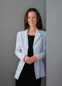 Corporate photo of Kate Schaffner. A woman in her thirties, she is standing and smiling. She has light skin and long brown hair. She is wearing a pale blue blazer and black dress.